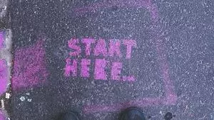 Two feet standing on a pavement in front of pink 'start here' graffiti