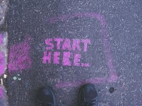 Two feet standing on a pavement in front of pink 'start here' graffiti
