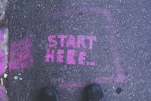 Two feet standing on a pavement in front of pink 'start here' graffiti